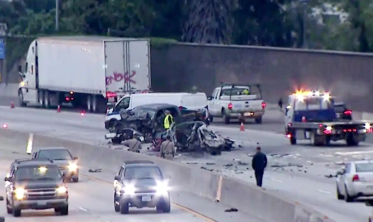 Damaged vehicles and emergency responders at a major freeway crash scene, with debris scattered across multiple lanes and traffic passing nearby.
